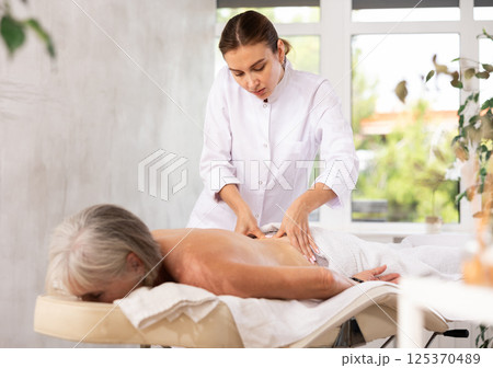 Young masseuse performing back massage to aged female client Young masseuse performing back massage to aged female client 125370489