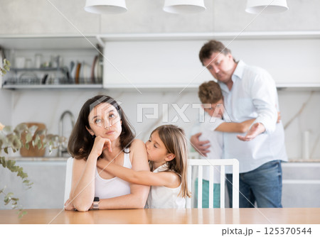 Offended mother sitting at the kitchen table being hugged by little daughter 125370544