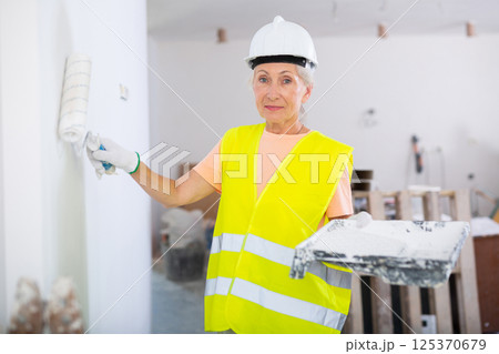 Portrait of elderly positive woman painter in protective helmet and yellow vest who paints walls with paint roller 125370679