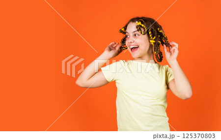 Styling tips. Small girl Curling Hair Using Curlers orange background. Adorable child hairdo. Teen hobbies. Daughter with curlers on her head laughing. Hairdresser salon. Female beauty routine 125370803