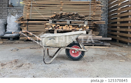 Wheelbarrow parked on a construction site with wood stacks in the background during daylight hours Wheelbarrow parked on a construction site with wood stacks in the background during daylight hours 125370845