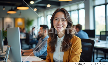 This photo depicts a young Asian woman in a professional office environment. She is sitting at a desk with a computer, wearing a white shirt and yellow blazer.  125371587