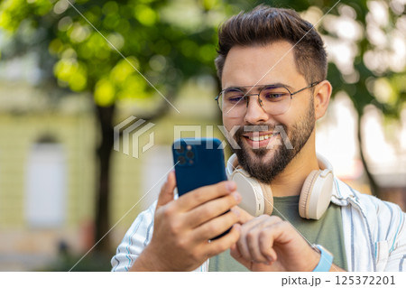 Smiling young man using smartphone typing texting scrolling social media app on city town street Smiling young man using smartphone typing texting scrolling social media app on city town street 125372201