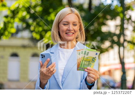 Caucasian mature businesswoman on city street showing earnings cash money with smartphone in hands Caucasian mature businesswoman on city street showing earnings cash money with smartphone in hands 125372207