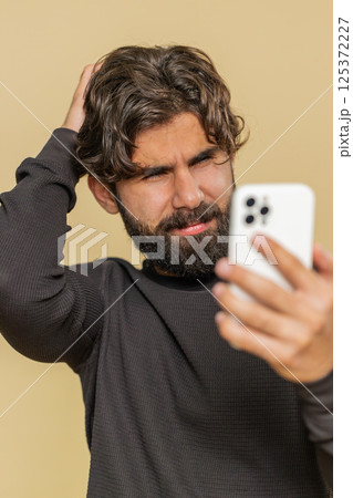 Upset Lebanese young man using mobile cell phone typing browsing, loses lottery on beige background Upset Lebanese young man using mobile cell phone typing browsing, loses lottery on beige background 125372227