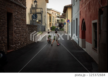 rear view of two small girls riding bicycle and scooter in the street of a town 125372884