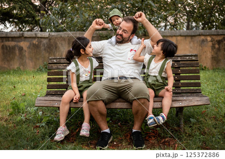 man sitting with small baby and two elder children on the bench in the park 125372886