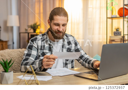 Young man checking bills at home table, laptop details, relief, calm smile, accurate finance match Young man checking bills at home table, laptop details, relief, calm smile, accurate finance match 125372937