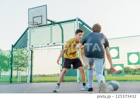 Two Young Caucasian male teenagers Playing Basketball at Park outdoor playground in sunset light. Teenagers' hobby, offline activity. Concept of sports, hobbies and healthy lifestyle. Selective focus. 125373432