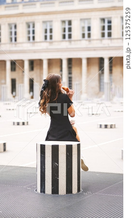 Back view of woman in black dress eating croissant in cozy French courtyard 125375259