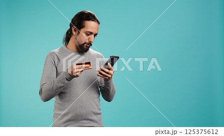 Elated man adding payment method on website, delighted to take advantage of promotions, studio background. Joyful person thrilled to do internet shopping, typing credit card data on phone, camera B 125375612