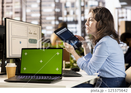 Woman employee examines business data next to mockup display, working hard at the office to meet deadlines and achieve objectives. Young adult studies infographics on computer. 125375642