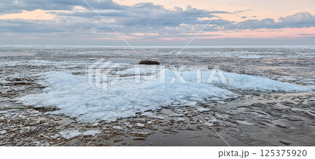 Sunrise over a tranquil coastline with icy patches and smooth stones along the sandy shore. The sky reflects in the water, creating a serene and peaceful atmosphere. 125375920