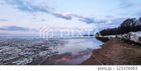 Spring landscape. Panoramic view of beautiful dawn on bay. Cumulus clouds over water in bright light. Ice, snow and rocks on coastline. Rising rays of sun are reflected in sea. High quality photo 125376063