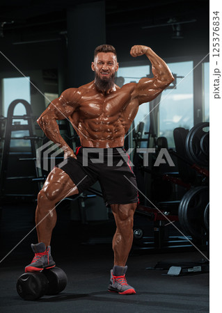 muscular individual poses proudly in fitness center emphasizing his sculpted physique. He stands on one leg showcasing his strength while surrounded gym equipment. 125376834