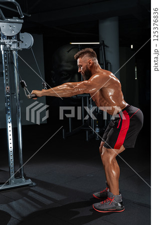 man muscular build focuses on his workout routine using cable machine in well equipped gym. lighting highlights his efforts and determination during exercise. 125376836