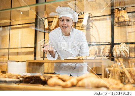Smiling young saleswoman in white chef jacket and cap standing behind bakery counter during working day in bakehouse. Cooking, culinary and bakery concept 125379120