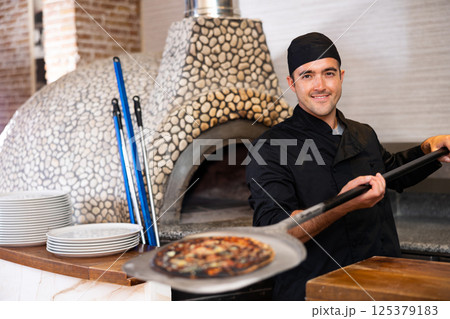 Latino chef in black uniform holding shovel with pizza in kitchen restaurant Latino chef in black uniform holding shovel with pizza in kitchen restaurant 125379183