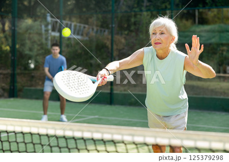 Sporty mature woman padel player hitting ball with a racket on hard court Sporty mature woman padel player hitting ball with a racket on hard court 125379298