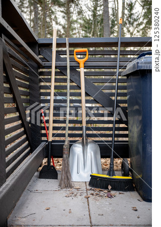 Various garden tools including broom, shovel, and hoe leaning against a wooden fence outdoors 125380240