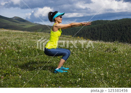 Woman doing squats at flowering grassland mountain top Woman doing squats at flowering grassland mountain top 125381456