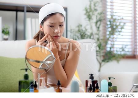 Young asian woman looking mirror with stress on facial sitting on sofa in living room at home. Young asian woman looking mirror with stress on facial sitting on sofa in living room at home. 125382170