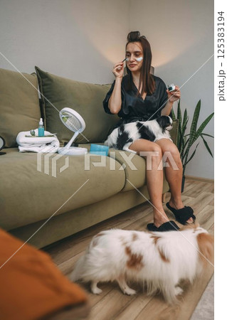 woman applying facial cream with applicator on green couch with japanese chin on her lap, another nearby on wooden floor, calm home routine, selfcare, pet companionship, lifestyle content 125383194