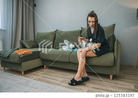 woman applying skincare lotion to her leg while sitting on green couch with two japanese chin dogs during relaxed morning beauty routine, pets calm, skincare, dog lovers, self care brands woman applying skincare lotion to her leg while sitting on green couch with two japanese chin dogs during relaxed morning beauty routine, pets calm, skincare, dog lovers, self care brands 125383197
