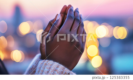 African young woman praying hands in warm sweater against city lights at dusk African young woman praying hands in warm sweater against city lights at dusk 125383355
