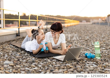 Young mother and daughter using laptop for online exercise session on rooftop, enjoying quality bonding and fitness. Family spends time healthily while performing fun Young mother and daughter using laptop for online exercise session on rooftop, enjoying quality bonding and fitness. Family spends time healthily while performing fun 125383524