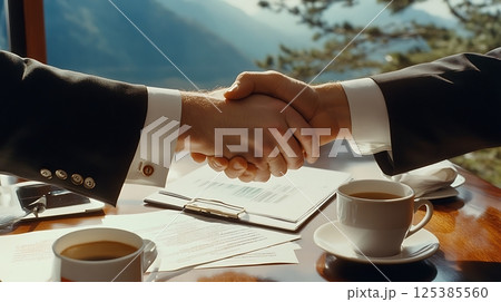 two businesspeople shaking hands after agreement in formal meeting setting, documents and coffee cups on table, 125385560