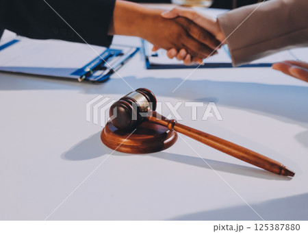 Two businesspeople are shaking hands in a law office with a scale and a gavel on the table 125387880