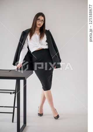 Confident businesswoman leaning on desk in office, wearing black suit, skirt and white shirt 125388677