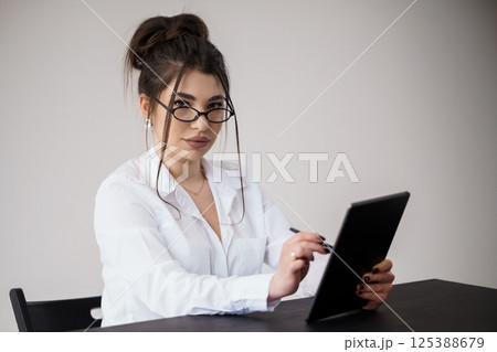 Young woman in white shirt using digital tablet at desk, focused expression, professional setting Young woman in white shirt using digital tablet at desk, focused expression, professional setting 125388679