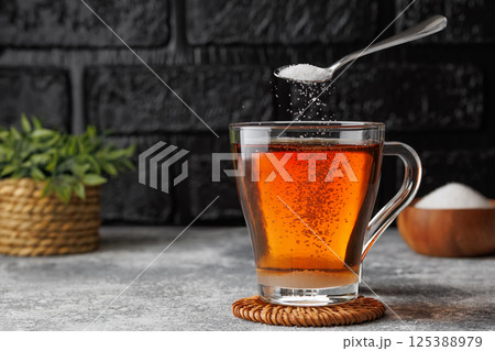 White sugar falling from teaspoon into glass cup of hot tea placed on coaster against black brick wall as background 125388979