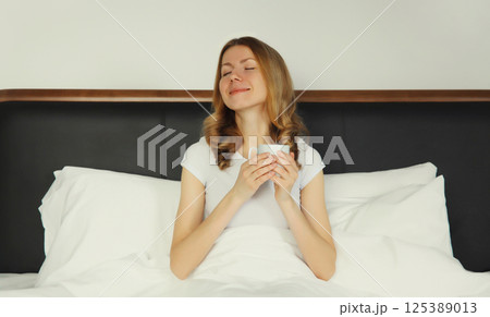 Happy smiling young woman enjoying with cup of hot coffee lying on bed at home 125389013