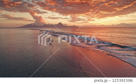Aerial view capturing waves gently crashing on the shore of Tahiti at sunset, reflecting vibrant orange and red hues amidst a dramatic cloudscape 125391506