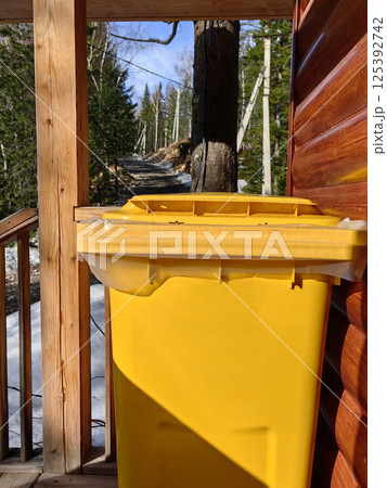 yellow trash bin placed near rustic cabin along forested path in bright daylight. close up. yellow trash bin placed near rustic cabin along forested path in bright daylight. close up. 125392742