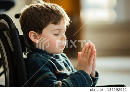 Young boy in a wheelchair with eyes closed and hands clasped in prayer, wearing a blue sweater, portraying faith, hope, and resilience 125392952