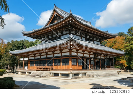 Ornate wooden Japanese temple with curved roof and intricate details, set against a blue sky with autumn trees in the background 125392956