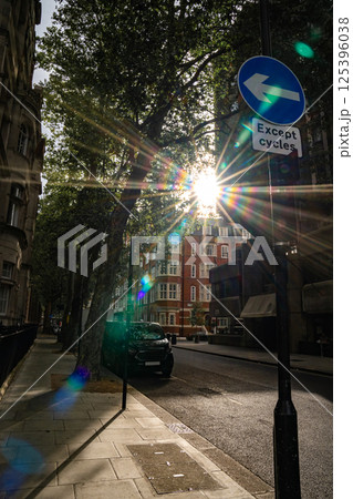 Sunlight shines through trees along a city street, with a road sign indicating a left turn for cycles. The urban scene features a quiet street with a parked vehicle and historic buildings in the Sunlight shines through trees along a city street, with a road sign indicating a left turn for cycles. The urban scene features a quiet street with a parked vehicle and historic buildings in the 125396038