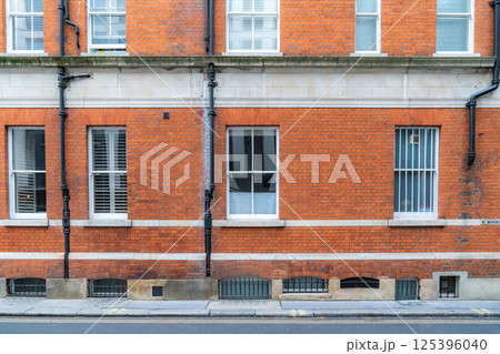 Red brick wall with a series of windows, pipes, and ventilation grills on a street in an urban setting. The building features a classic design with a solid structure and clear architectural lines. Red brick wall with a series of windows, pipes, and ventilation grills on a street in an urban setting. The building features a classic design with a solid structure and clear architectural lines. 125396040