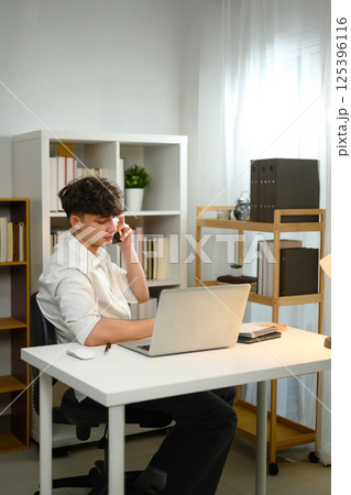 Smiling entrepreneur holding smartphone conversation while seated at desk with laptop 125396116