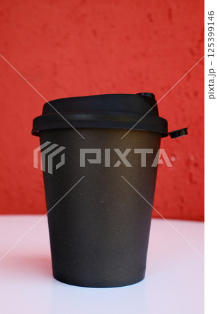Black disposable coffee cup with lid on white table against vibrant red textured wall background 125399146
