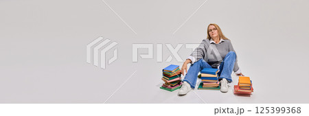 Middle-aged woman sitting on floor surrounded by piles of books with tired expression. 125399368