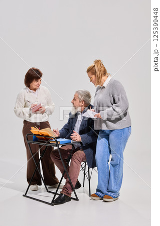 Three elderly students, man and women with books and notes interacting during classroom activity against white studio background. 125399448