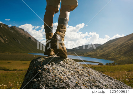 Hiker legs wearing leather boots hiking on high altitude mountain top,with a lake in the distance 125400634