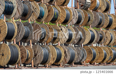 Wide view of storage of thick black industrial cable tightly coiled on wood reel at outdoor storage. Wide view of storage of thick black industrial cable tightly coiled on wood reel at outdoor storage. 125401666