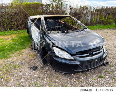 A completely burned-out black sedan with extensive fire and impact damage sits abandoned on a rocky patch beside a hedgerow in a rural area A completely burned-out black sedan with extensive fire and impact damage sits abandoned on a rocky patch beside a hedgerow in a rural area 125402035