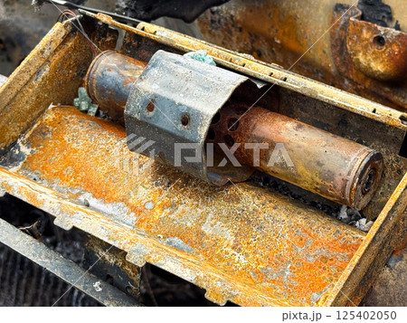 Burned steering column lock from a car with heavy rust and fire damage, showing the metal locking mechanism and oxidized surface inside a charred dashboard frame 125402050